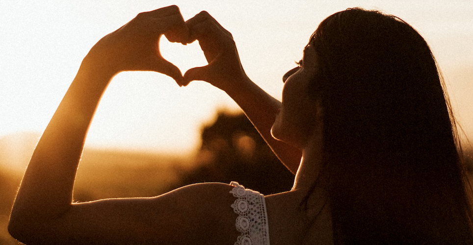 woman in silhouette holding hands in shape of a heart up to a sunrise.