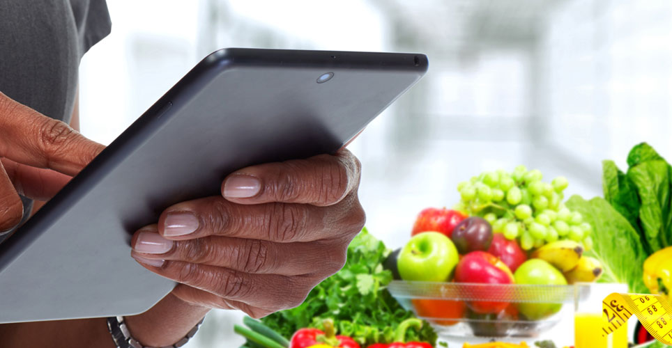 close up of an African-American hand holding a tablet with images of fruit and a sewing tape measure in the background.