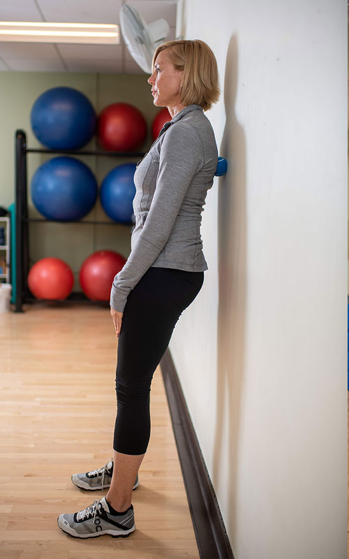 woman standing against wall with a small ball between her upper back and wall showing erector spinae start movement