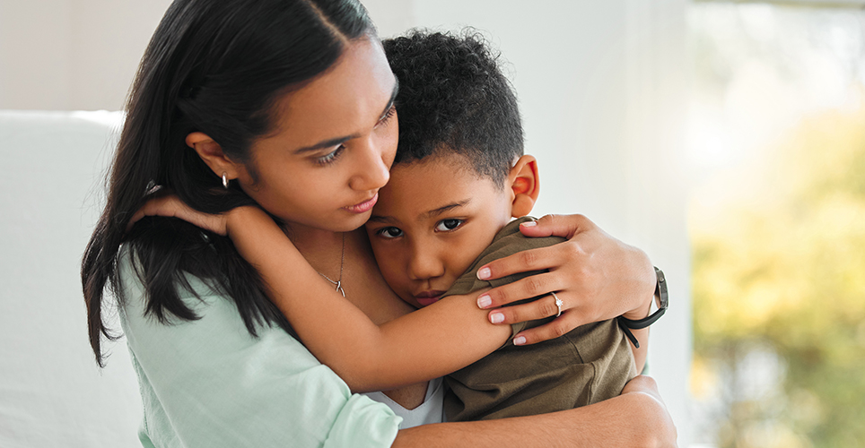 Female caregiver comforting a child with a hug. Both have darker skin and hair.