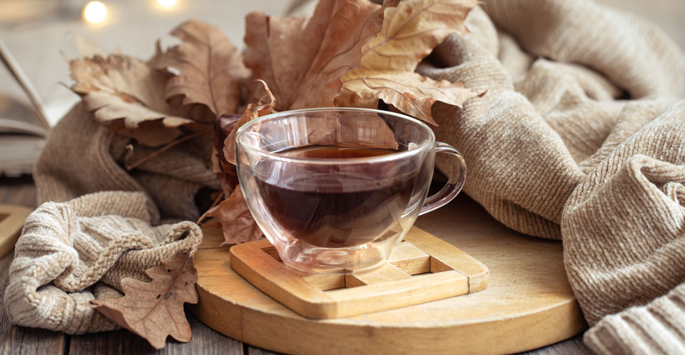 Close up of a clear tea cup filled with tea surrounded by a cream sweater and rust-colored leaves