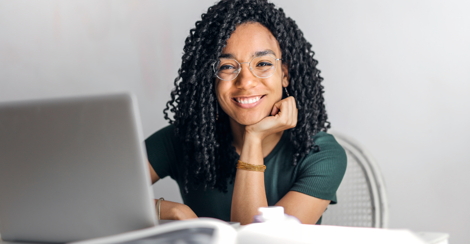 Person sitting at table resting chin on hand and smiling while laptop is open.