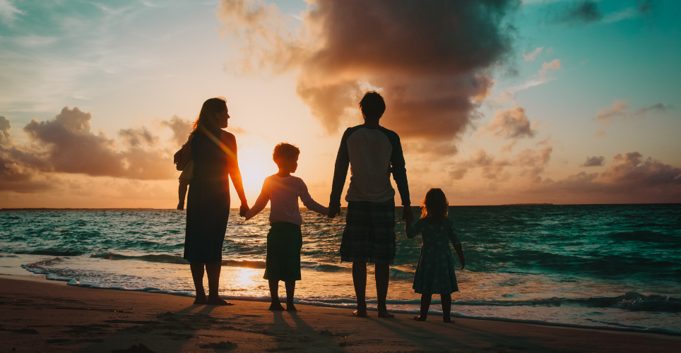 Family holding hands on beach, facing away from camera silhouetted by sunset
