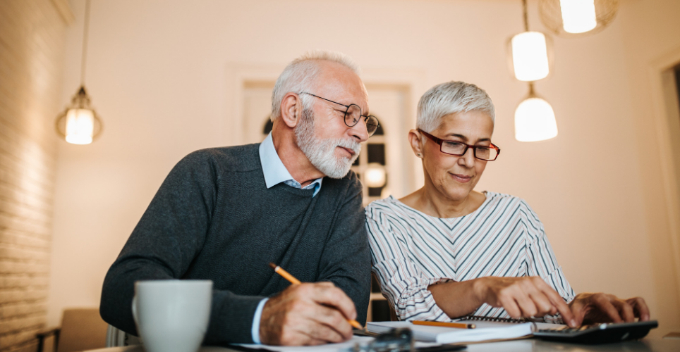 A retirement-aged man and woman sitting at a table, planning, with paper, pens and calculator.