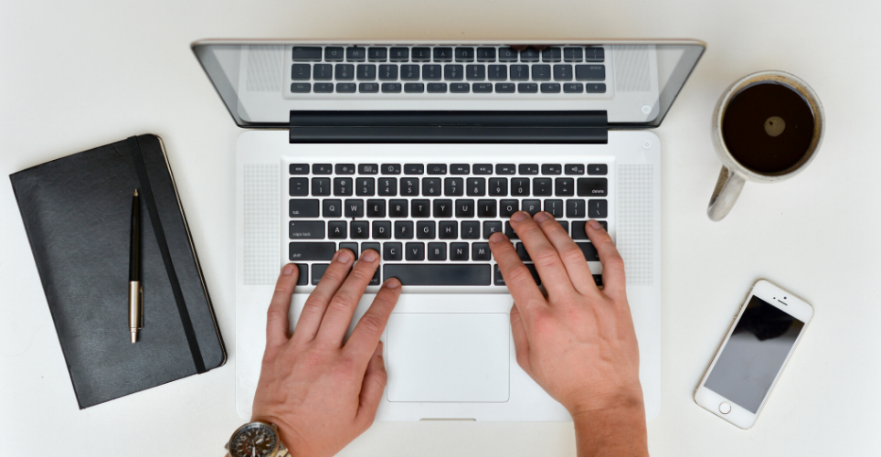 Close up of two hands typing on a lap top keyboard with a cellphone and cup of coffee