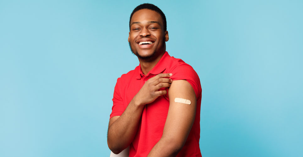 Waist-up shot of smiling African-American man in red shirt showing off a bandage that denotes he was vaccinated.