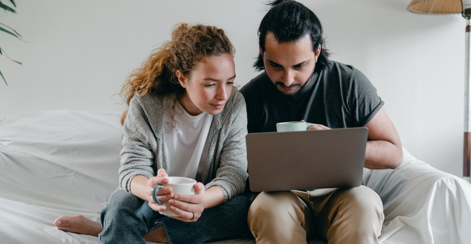 Two people sitting on a coach, holding coffee cups and looking at a laptop screen.